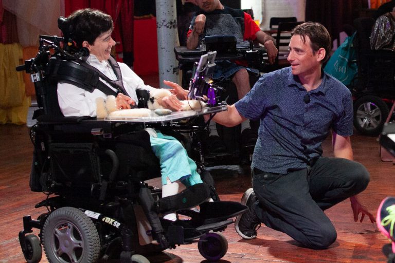 man in blue shirt crouches and smiles at woman in white shirt sitting in wheelchair who smiles back