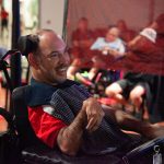 man in red shirt and striped blue scarf sitting in wheelchair smiles