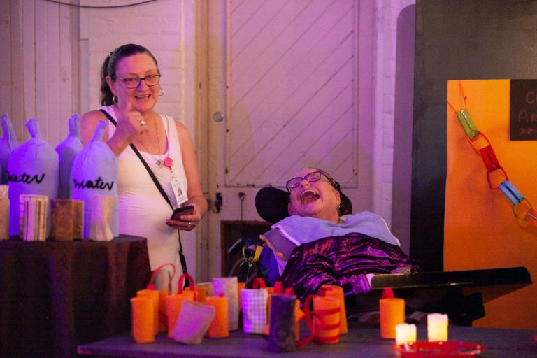 woman in white tank raises her hand with a thumbs up while woman in purple sequin jacket sitting in wheelchair looks over and laughs behind bar with fabric props