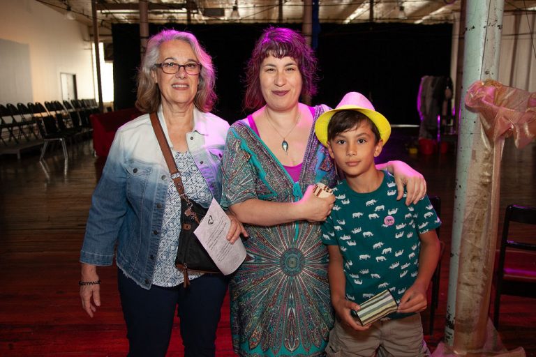 Woman in ombre jean jacket poses and smiles with younger woman in blue patterned dress and young boy in yellow hat